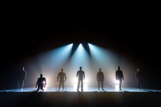 Ill-Abilities band standing one by one on stage, illuminated by spotlights from behind.