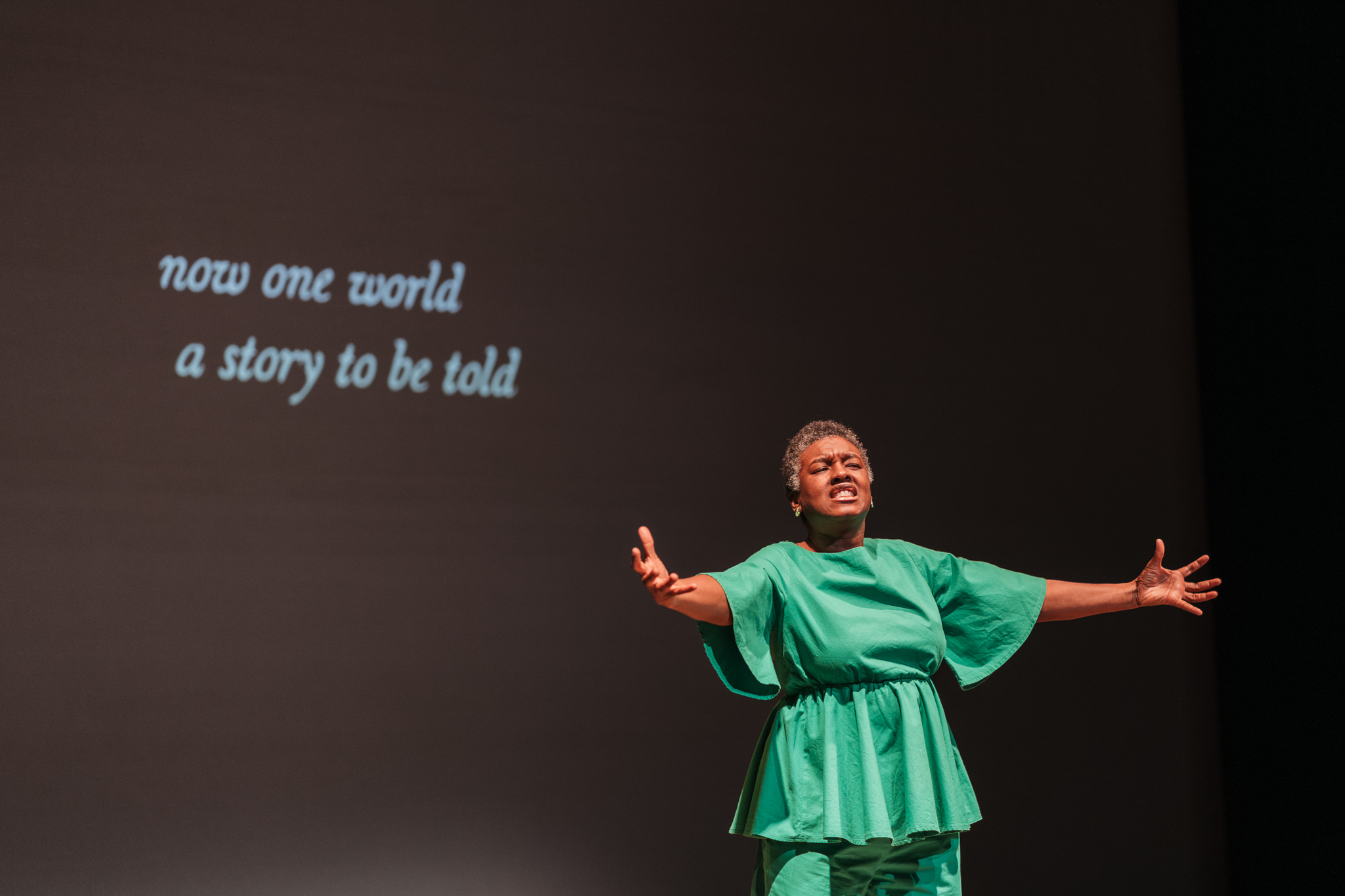 Image Description: Jo-Anne, a Black Bajan woman with a short fro, wearing a bright green shirt and pants, is standing on stage with her arms open. Behind her, white font on a black background reads "now one world, a story to be told." Photo by Lindsey Tran.