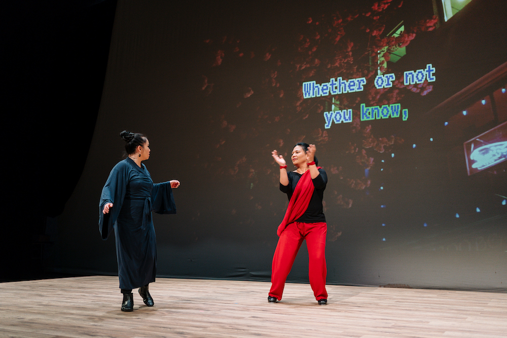 Image Description: Carmelle, Filipina woman with long black hair, wearing black shirt, red pants and a sash, and King, a Vietnamese woman with black hair tied in a high bun, wearing a dark blue robe are performing a song and dance on stage. Behind them, blue karaoke font against a blurred pink cherry blossom tree background reads "whether or not you know." Photo by Lindsey Tran.