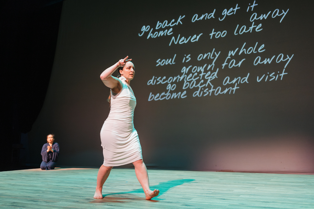 Image Description: Jordan, a white woman with long brown hair in a ponytail, wearing a white, Grecian-style dress, and King, a Vietnamese woman with black hair tied in a high bun, wearing a dark blue robe are on stage in a song and dance. Jordan is in the foreground, mid-walk with her right hand gesturing forwards in the "see" handshape. King is in the background, kneeled, hands in the shape of a book. Behind them, white text on a black background reads "go back and get it, home, away, never too late, soul is only whole, grown far away, disconnected, go back and visit, become distant." Photo by Lindsey Tran.